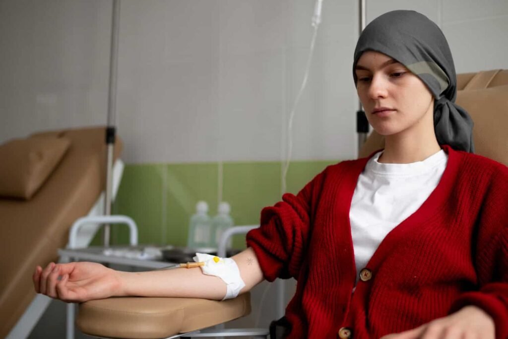Female patient receiving a blood test for cancer screening, wearing a red cardigan and headscarf, with IV setup and medical equipment in a clinical setting.